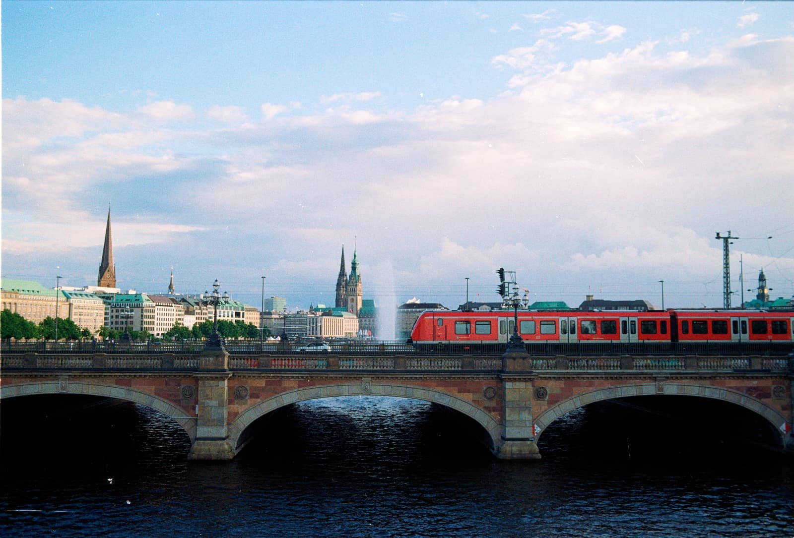 De Lombardsbr&uuml;cke met de skyline van Hamburg op de achtergrond en een rode trein op de brug, gezien vanaf de Kennedybr&uuml;cke